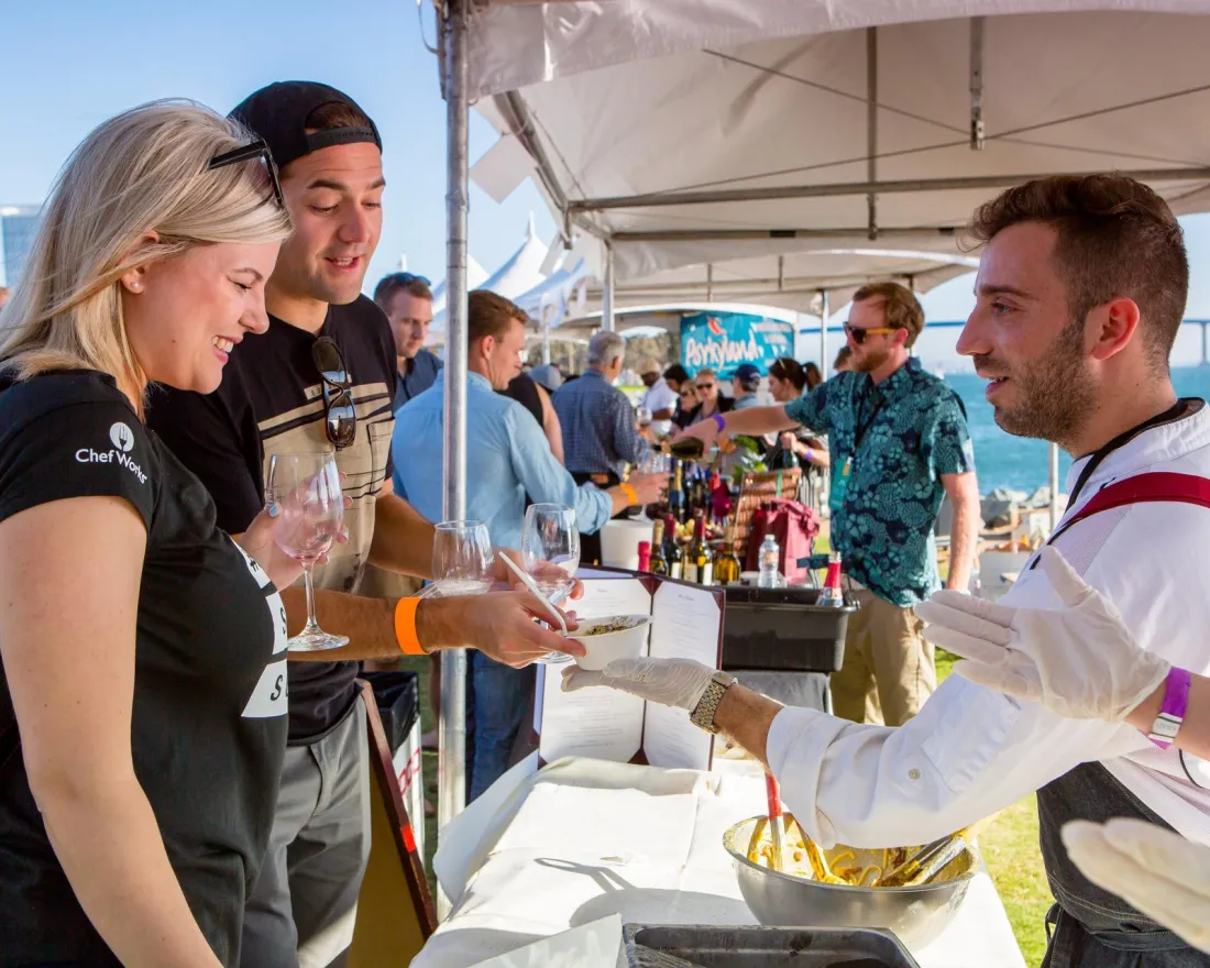 A food vendor serves dishes to two smiling guests at a bustling outdoor festival in San Diego, CA