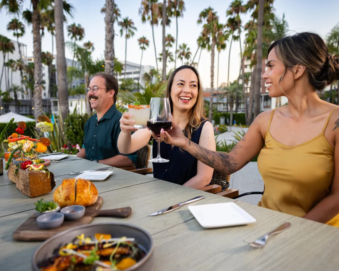Three friends enjoying a meal outdoors, toasting with drinks amidst palm trees in San Diego, CA.