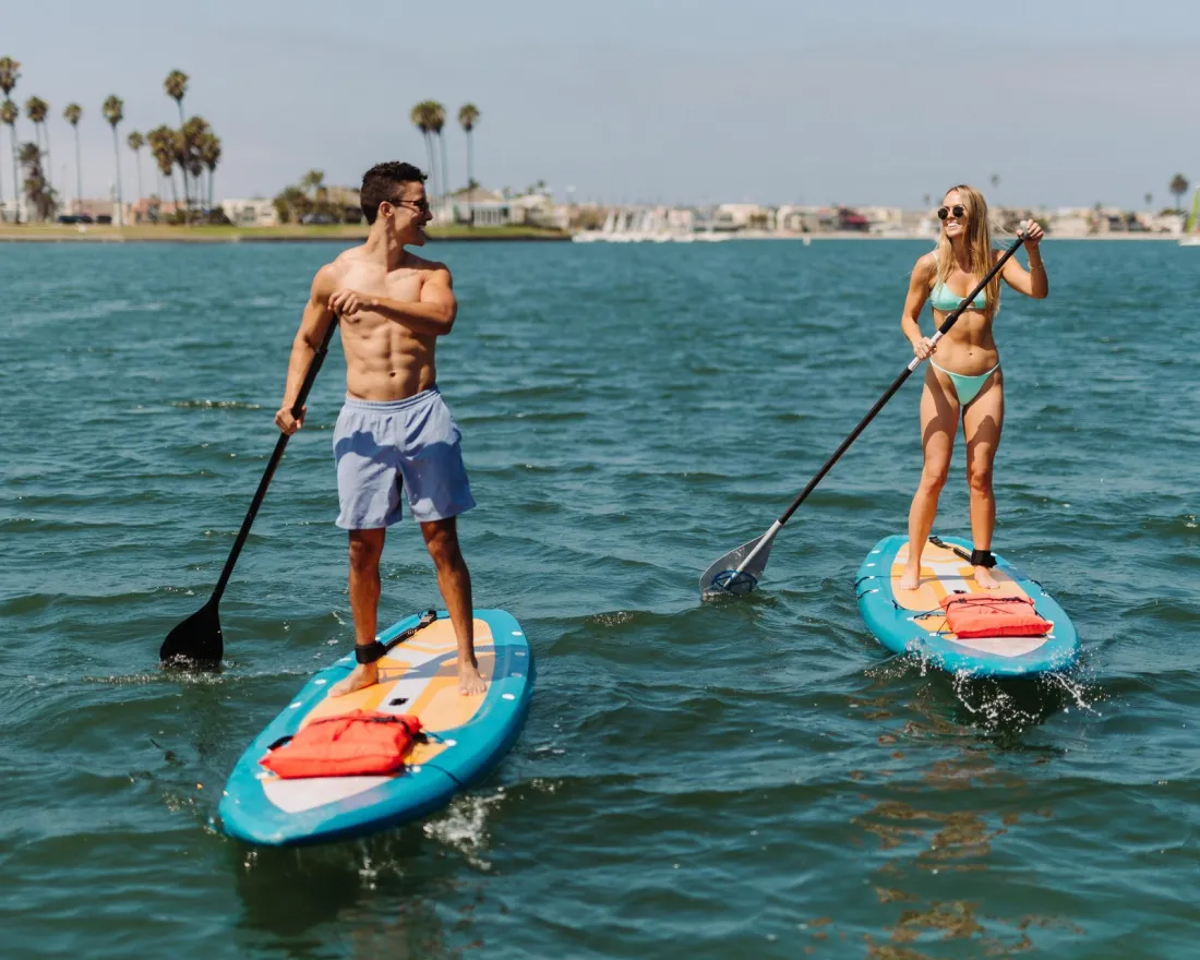 Two individuals paddleboarding on Mission Bay in San Diego, surrounded by palm trees and a distant marina. The sun shines brightly overhead.