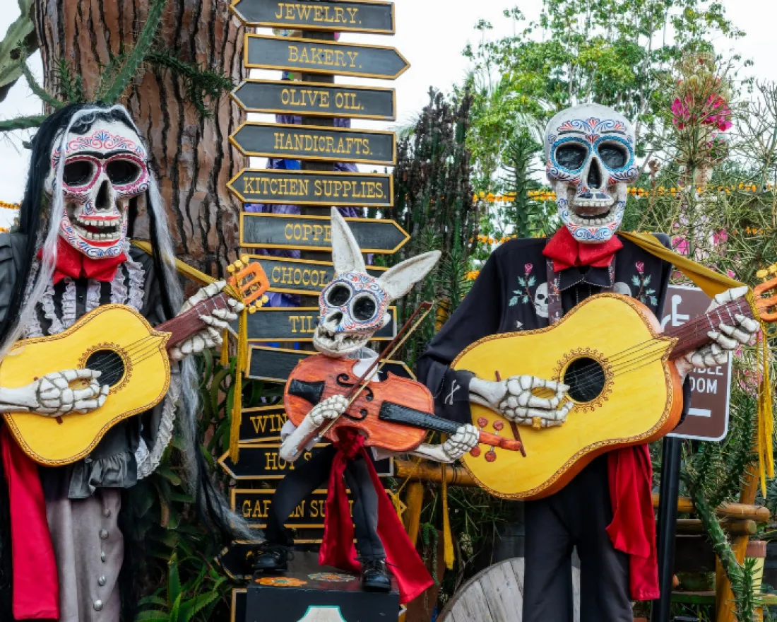 Three skeleton figures playing guitars, surrounded by greenery and colorful signage.