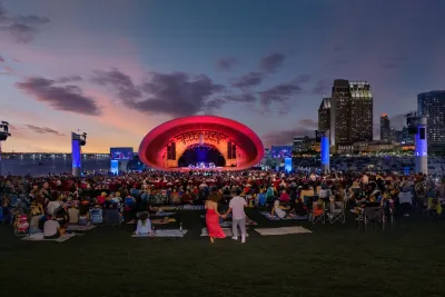 A large crowd gathers on a lawn facing an outdoor concert stage illuminated in red, with city buildings and a sunset sky in the background.