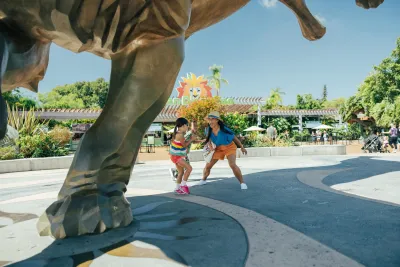 Two people pose playfully under a large dinosaur statue at a zoo or amusement park on a sunny day.