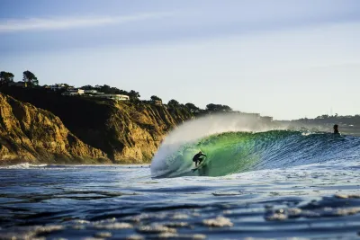 A surfer rides inside the barrel of a wave near a rocky coastline with cliffs and houses visible in the background.