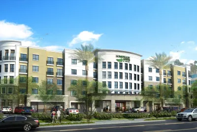 A modern four-story Homewood Suites hotel in San Diego, with green and beige exterior, large windows, and palm trees, with cars parked in front and people walking along the sidewalk.