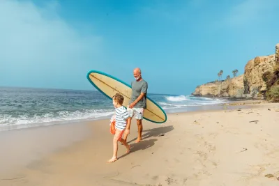 An adult and a child walk barefoot on a sandy beach; the adult carries a surfboard while waves approach the shore in the background.