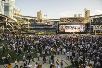 A large crowd gathers on a baseball field in front of a big screen during an event at a stadium, with city buildings visible in the background.