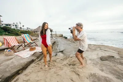 A man takes a photo of a woman in a red swimsuit on a sandy beach near picnic chairs, a blanket, and an ocean backdrop.