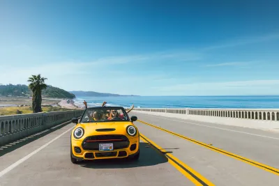 A yellow convertible with its top down drives on a coastal highway bridge next to the ocean in Torrey Pines under a clear blue sky, with passengers raising their arms.