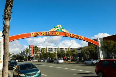 A large red arch with "Barrio Logan" written on it spans a street lined with parked cars and buildings under a blue sky.