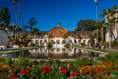 Balboa Park Botanical building with arched windows behind a reflective pond, surrounded by colorful flower beds, palm trees, and visitors on a clear day.