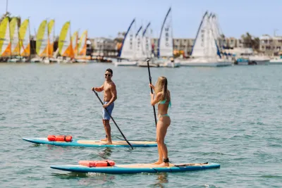 A man and a woman stand-up paddleboarding on calm waters of Mission Bay, with sailboats and buildings visible in the background.