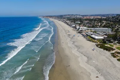 Aerial view of Del Mar's wide sandy beach with waves along the shoreline, people scattered on the sand, and buildings lining the coast under a clear blue sky.