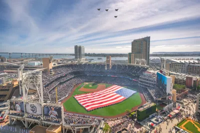 A large American flag is displayed on petco park, with four jets flying in formation overhead and a crowd filling the stands.