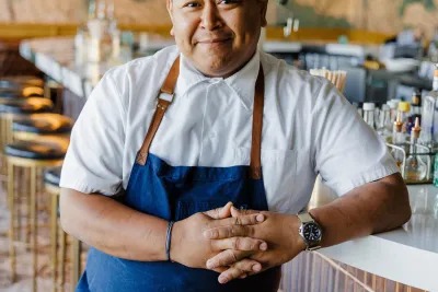 A chef in a white shirt and blue apron, David Sim, stands behind a bar counter, smiling with arms folded, in a modern restaurant setting.