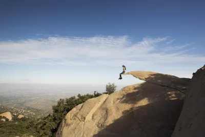 A person sits on the edge of a large rocky outcrop above a landscape, with a clear sky and distant hills visible in the background.