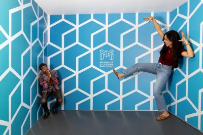 Two people pose in an optical illusion room at the Museum of Illusions San Diego; one appears large and the other small due to the room’s angled walls and geometric blue pattern.