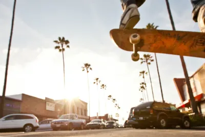 A skateboarder performs a trick mid-air above a city street lined with cars, palm trees, and buildings on a sunny day.