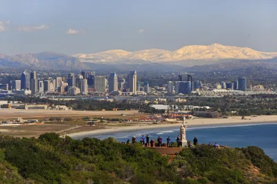 A group of people stands on a hill overlooking the San Diego skyline, with a beach, bay, and distant snow-capped mountains in the background.