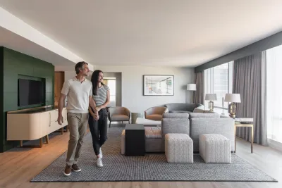 A man and woman walk together and smile inside a modern, spacious living room with neutral decor, large windows, and contemporary furniture.