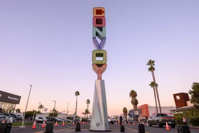 A tall vertical sign spelling "Convoy" in colorful letters stands in the middle of a street lined with palm trees, cars, and shops at sunset.