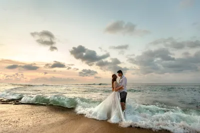 A bride and groom stand together at the shoreline, embracing in the surf at sunset with clouds scattered across the sky.