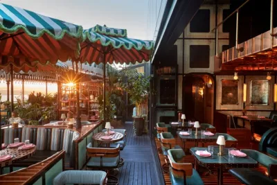 Outdoor restaurant with striped awnings, round tables set with pink napkins, cushioned seating, and potted plants, with sunlight streaming in from the left.
