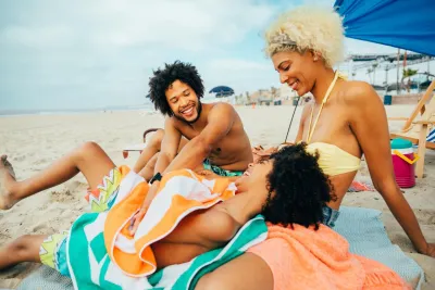 Three people sit smiling and laughing together on a beach, sitting on towels under a canopy with sand and ocean visible in the background.