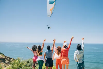 Six people stand on a grassy bluff at the Torrey Pines Gliderport in the La Jolla neighborhood of San Diego, facing the ocean with arms raised as they watch a paraglider flying over the water under a clear blue sky.
