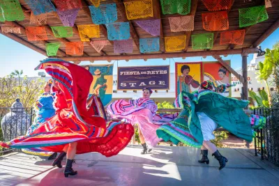 Three dancers in colorful traditional dresses perform on an outdoor stage at Fiesta de Reyes in San Diego's Old Town, decorated with papel picado banners and Fiesta de Reyes signs in the background.