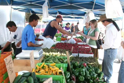 People shopping for fresh vegetables and produce at a farmers market stand in the Hillcrest neighborhood of San Diego, with greens, cucumbers, peppers, and other vegetables displayed on tables under a canopy.