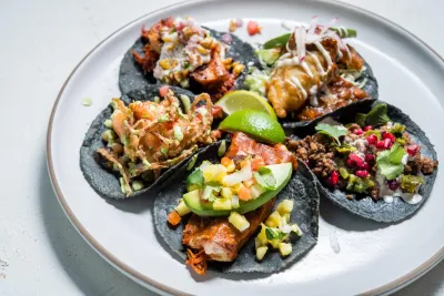 A plate with five assorted tacos from Puesto on blue corn tortillas, topped with various meats, vegetables, sauces, and garnishes, with lime wedges in the center.