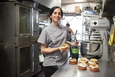 Chef Vivian Hernandez of Azucar, in a gray uniform, stands in a commercial kitchen, holding a tart and a whip cream canister, with several finished tarts on the counter beside her.