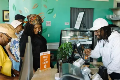 Customers wait at the counter at The Mental Bar in San Diego while a staff member in a white hoodie and cap assists them in a brightly decorated cafe with a "Pick Up Here" sign.