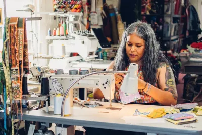 Claudia Rodriguez Biezunski is sewing fabric at a worktable in the colorful Sew Loka sewing studio with various supplies and a sewing machine.