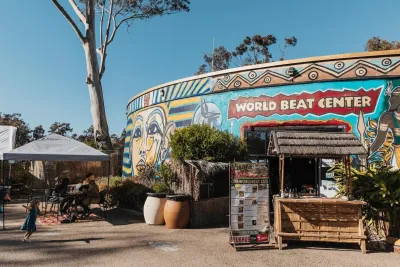 Outdoor view of the WorldBeat Center in Balboa Park, featuring a colorful mural, a small café stand, umbrellas, and people sitting and walking in the sun.