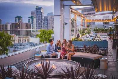 Four people sit around a fire pit on a rooftop lounge at sunset, surrounded by modern buildings and string lights, with city views in the background.