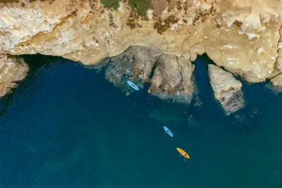 Aerial view of three paddleboards, one orange and two blue, floating near rocky cliffs in clear blue water.