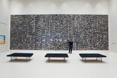 A person stands in front of a large wall display filled with many small framed photographs in a spacious, modern gallery with three black benches.
