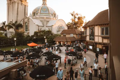 People gather in an outdoor courtyard with tables and umbrellas near historic buildings and a domed structure under a clear sky.