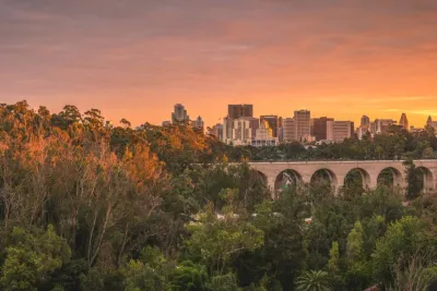 San Diego skyline at sunset with trees in the foreground, a historic viaduct, and an airplane flying in the orange sky.