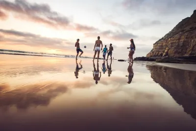 A group of people stand and play with a ball on a wet sandy beach at sunset, with cliffs and ocean visible in the background.