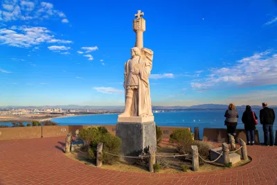 A stone statue of a historical figure stands on a raised platform overlooking the ocean, with people observing the coastal view and city skyline in the background.
