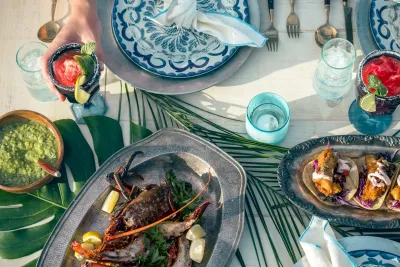 A table set with seafood, tacos, guacamole, blue and white plates, drinks with ice and lime, and cutlery, seen from above. A hand reaches for a red cocktail.