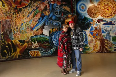 Two people stand closely together in front of a large, colorful mural featuring abstract and cultural imagery.