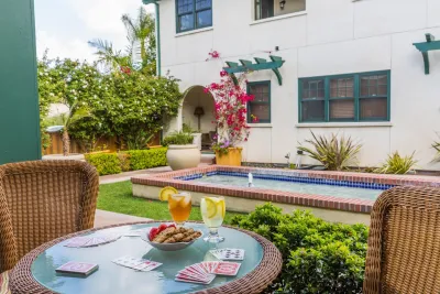 A round wicker table with playing cards, cookies, drinks, and fruit sits on a patio overlooking a small pool and garden next to a white building.