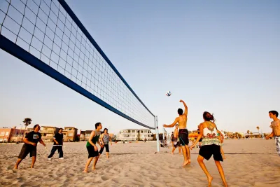 A group of people play beach volleyball on sand near the ocean, with houses and palm trees visible in the background under a clear sky.