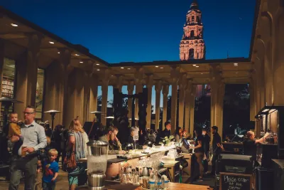 People gather at an outdoor bar and dining area in the evening; a tall illuminated tower is visible in the background against a dark blue sky.