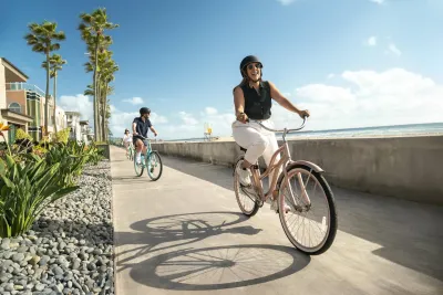 Two people ride bicycles along a beachside path on a sunny day, with palm trees, buildings, and the ocean visible in the background.