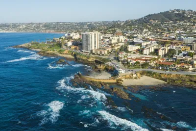 A coastal city with buildings along the shoreline, rocky cliffs, and waves crashing onto the beach under a clear blue sky.