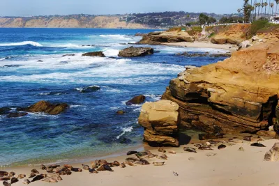 Rocky shoreline with waves crashing, sea lions resting on the sandy beach, and cliffs in the background under a clear sky.